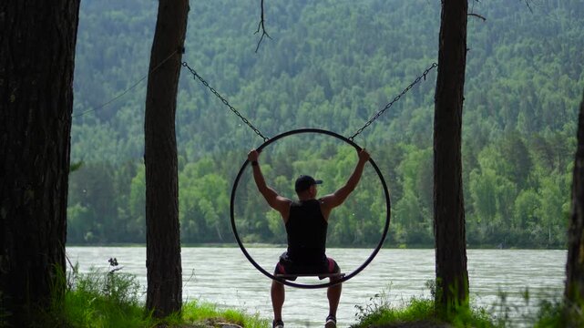 Inspiring static footage from a back view of a male traveler sitting in a circular hanging swing, then lifting his arms to hold the metal frame over the Katun River in Altai, Siberia.