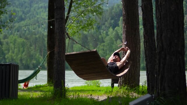 Inspiring static footage of a relaxed male traveler lying in a wooden hammock, stretching his arms overhead and then placing them behind his head on the Katun River bank in Altai, Siberia.