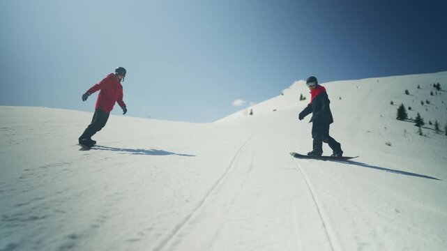 Two people snowboard down a snow-covered mountain slope. They enjoy winter sports on a clear, sunny day.