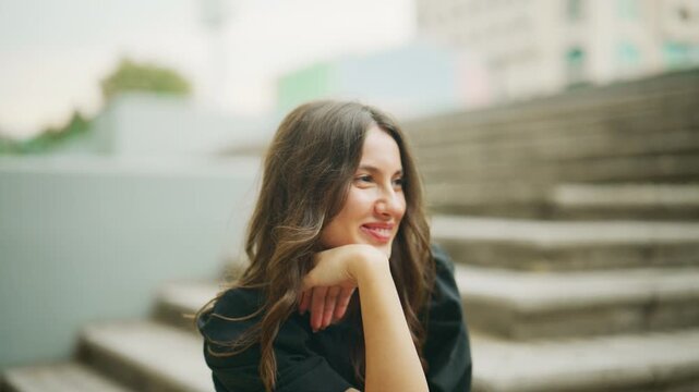 A happy young woman with a beautiful smile expresses joy and laughter in an urban outdoor setting. She sits relaxed on steps, showcasing positive emotions.