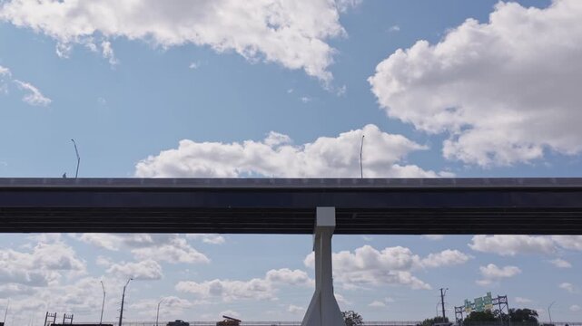 Aerial moving dolly shot of highway interstate 70 underpass looking up beneath multilane roadway with white fluffy clouds and summer sunny sun as concrete traffic structure stands tall