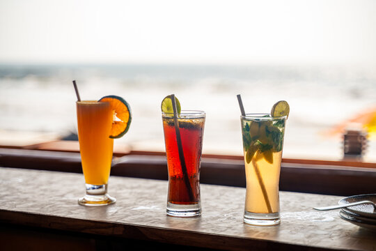 Various flavoured three glasses of mocktail with straws in it kept on the wooden table at beach side shacks. Beach and sea at background blur during early evening time.