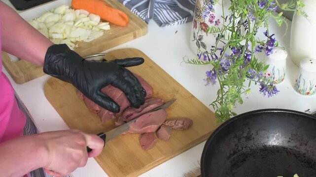 A woman in a kitchen cuts beef liver into pieces for frying. Nearby, there are vegetables on a cutting board and flowers in a vase on the table.
