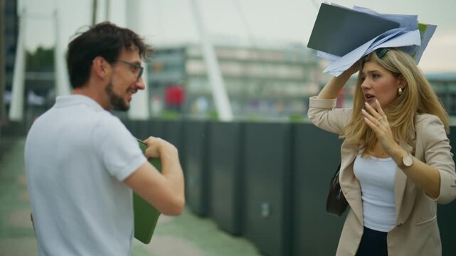 Two business colleagues encounter strong winds, causing their papers to fly while walking outdoors. The man and woman try to manage their documents and the challenging weather conditions.