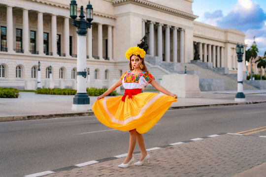 Latina woman celebrating Day of the Dead with traditional Mexican Catrina makeup and a vibrant dress, posing outdoors against the backdrop of the iconic El Capitolio building in Havana