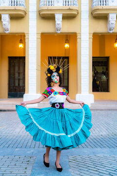 Latina woman with colorful skull makeup and a flower headdress, wearing a traditional embroidered blouse and flowing turquoise skirt, posing in front of a colonial building in Havana, Cuba