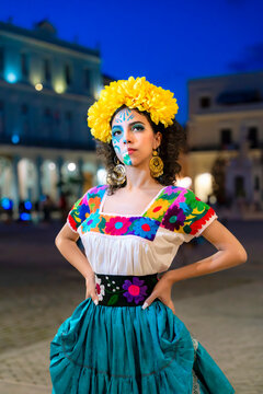 Young Latina Catrina in colorful embroidered dress and floral crown, skull makeup glowing at twilight in Havana, posing outdoors with vibrant traditional Day of the Dead style