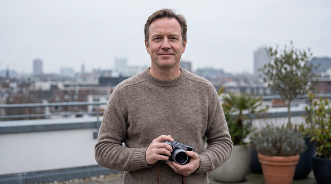 Mature man holding camera on rooftop terrace with distant city skyline behind