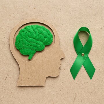 Green brain model inside human head cutout next to green awareness ribbon on cork