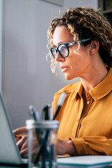A focused woman wearing glasses works at a laptop in a bright home office, typing and reviewing...