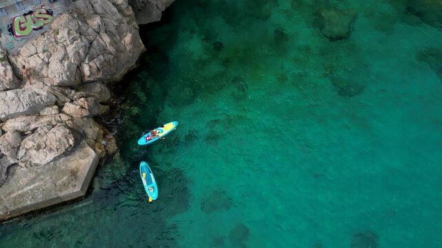 Top down aerial drone view of two people stand up paddleboarding in crystal clear turquoise sea near rocky cliffs at Plage des Pissarelles nudist cove, Cap d'Ail, French Riviera 