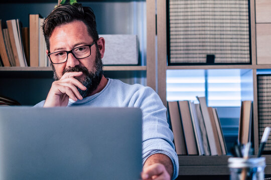 A thoughtful bearded man wearing glasses works on a laptop in a cozy home office, surrounded by books and shelves. He appears focused and contemplative while planning or researching