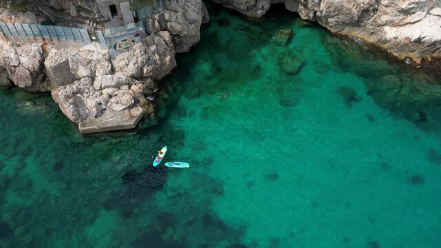 Top down aerial drone view of two people stand up paddleboarding in crystal clear turquoise sea near rocky cliffs at Plage des Pissarelles nudist cove, Cap d'Ail, French Riviera 