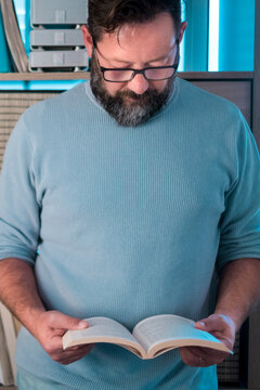 A bearded man wearing glasses reads an open paperback book in a cozy home setting, focused and relaxed. He wears a light blue sweater, standing in front of shelves with storage boxes,