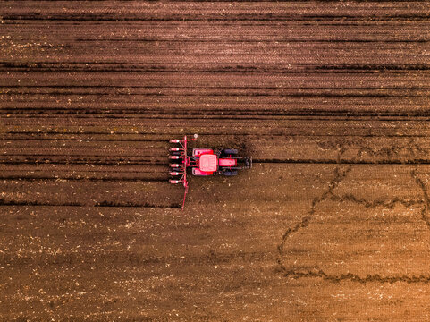 Red tractor pulling a seeder, planting seeds in a cultivated brown soil field, showing modern agricultural work from above