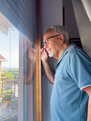 Fototapeta premium Elderly man looking through a window with reflection, thoughtful and contemplative at home on a balcony view