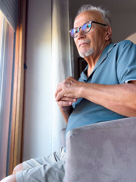 Senior man with glasses sitting by a window, looking contemplative and relaxed while enjoying natural light and quiet reflection