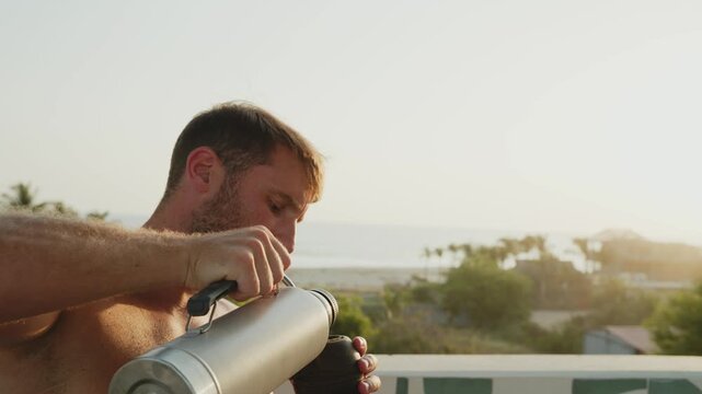 Shirtless man enjoying a traditional yerba mate drink on a balcony overlooking the ocean at sunset