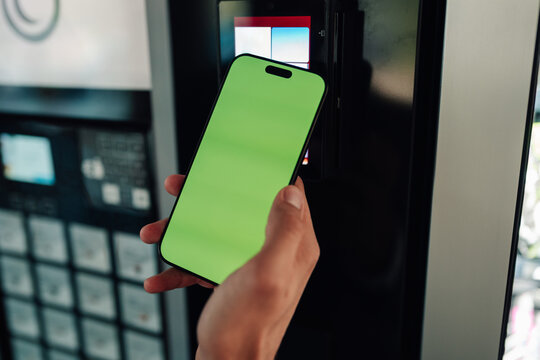 Hand holding smartphone with blank green screen, making a contactless payment at an automated vending machine