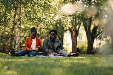 Fototapeta premium Happy black couple enjoying a picnic lunch in a sunny park, eating healthy food outdoors together
