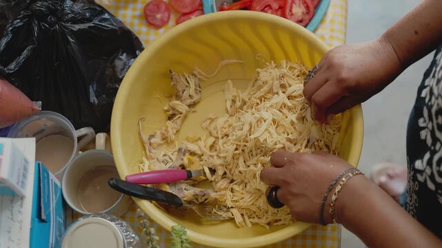 Top down view of a woman's hands shredding chicken meat in a yellow bowl to prepare a meal tinga
