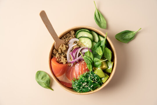 Healthy protein bowl with quinoa, smoked salmon, spinach, cucumber, avocado and chuka seaweed salad in a cardboard container on a light beige background