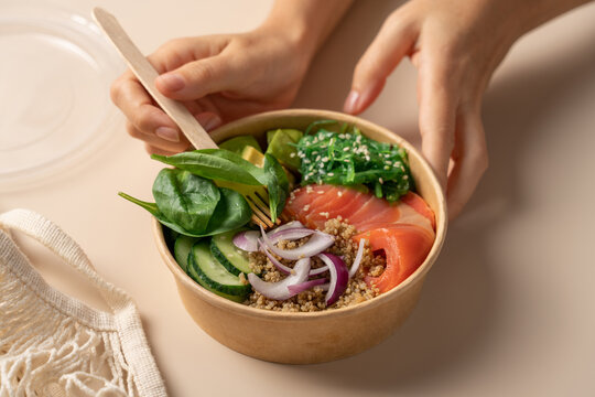 Hands holding a healthy protein bowl with quinoa, smoked salmon, spinach, cucumber, avocado and chuka seaweed salad in a cardboard container on a light beige background