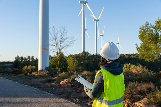 Engineer wearing a hard hat and safety vest standing at a wind farm, observing turbines and holding a clipboard