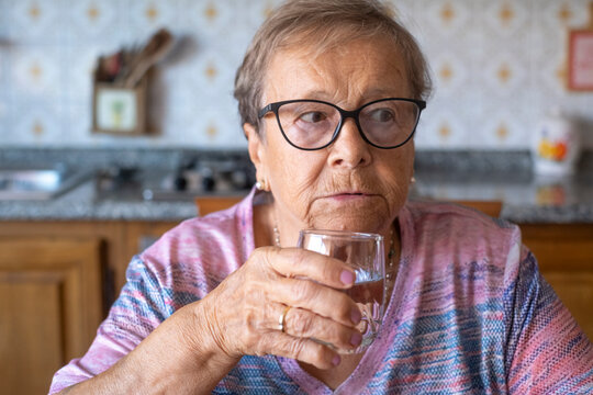 An elderly woman wearing glasses sits at a kitchen table, thoughtfully sipping a glass of water while looking to the side. The candid portrait captures quiet reflection, daily routine,