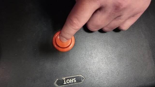 A hand pressing an orange button labeled 'SHOT' on a black surface in a close-up view