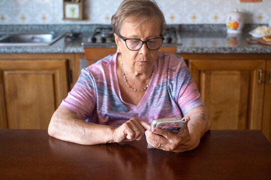 Elderly woman with glasses sits at a kitchen table concentrating on her smartphone, tapping the screen with focus. A senior grandmother practices digital skills, connecting with family