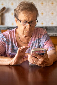 An elderly grandmother concentrates as she uses a smartphone at the kitchen table, tapping the screen with careful fingers. Wearing glasses and a colorful knit top, she practices digital communication