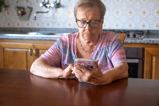 An elderly woman sits at a kitchen table focused on her smartphone, tapping the screen while wearing glasses and a casual knit top, conveying concentration, solitude and modern connectivity 