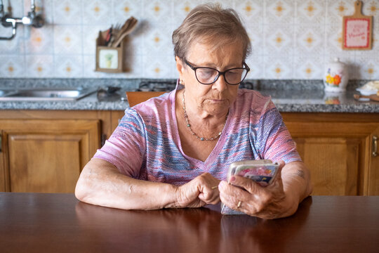 An elderly woman sits at a kitchen table concentrating on her smartphone, learning or messaging and staying connected with family while showing independence