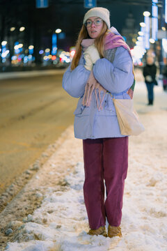 Young woman bundled in hat, scarf and gloves waits alone at a snowy city bus stop after sunset, standing on icy pavement under blurred streetlights in freezing winter air