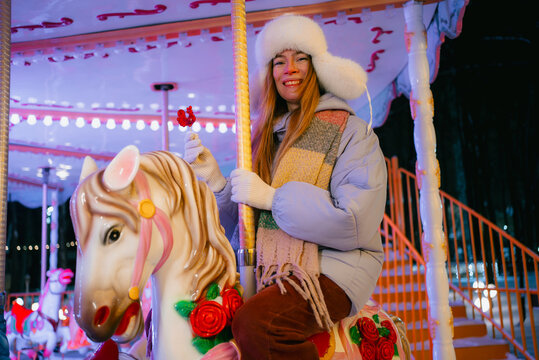 Woman enjoying a winter night carnival ride, smiling broadly while sitting on a carousel horse, experiencing joy and childhood nostalgia in an illuminated amusement park