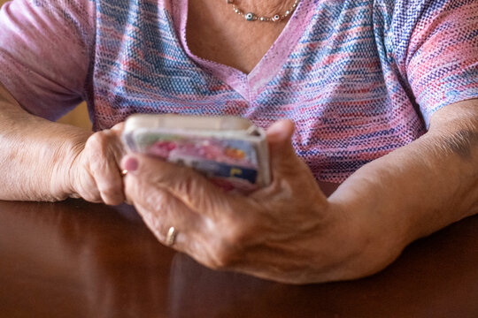 An older woman sits at a wooden table holding a smartphone, focused on the touchscreen while wearing a pink patterned shirt and delicate necklace. The close-up of her hands 