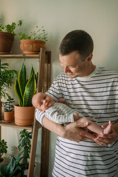 Young father tenderly holding his newborn baby, looking down and smiling, both relaxing at home surrounded by green houseplants