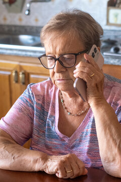 Elderly woman talking on smartphone at kitchen table looking thoughtful and concerned while wearing glasses and a patterned shirt