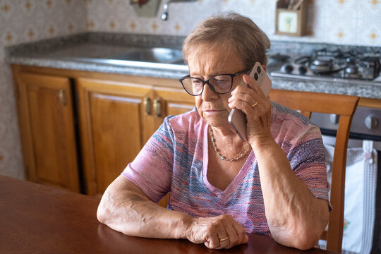 An elderly woman sits at a kitchen table speaking on her smartphone with a concerned expression, listening intently during a phone call that suggests worry about family or health 