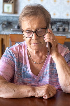 Elderly woman talking on a smartphone at home, a thoughtful senior making a phone call while seated at a kitchen table, wearing glasses and casual clothes with a quiet, reflective expression