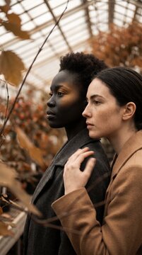 Two Women With Diverse Skin Tones Stand Together In A Quiet Autumn Greenhouse
