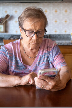 An elderly woman wearing glasses concentrates on her smartphone while sitting at a kitchen table, quietly connecting with family and technology in a domestic, thoughtful moment