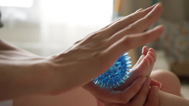 Careful mother or therapist giving a gentle foot massage to a newborn baby with a special blue spiky ball, a health procedure for infant wellness and muscle tone development