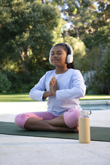 Fototapeta premium African American child meditating on yoga mat poolside in pink leggings with headphones and bottle