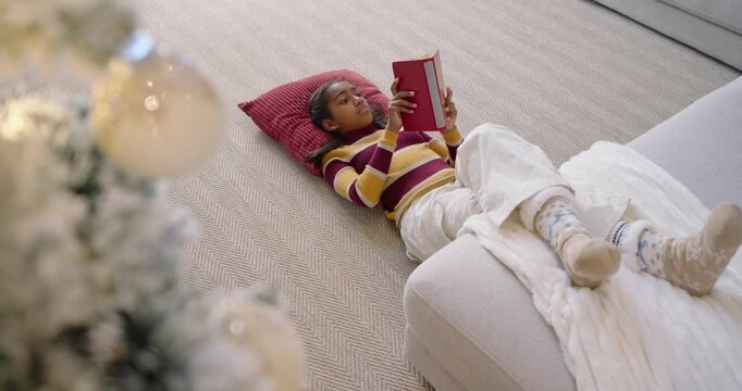 African American teen wearing striped sweater settling on carpet by sofa, opening book and studying