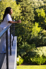 African woman leaning on metal rail holding mug in loungewear on balcony facing garden, copy space © WavebreakMediaMicro