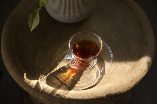 Clear glass teacup on saucer is sitting on textured table as sunlight is casting refracted patterns
