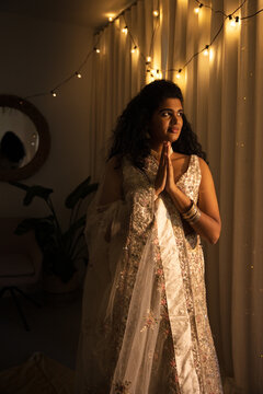 Indian adult woman standing pressing hands in namaste in cream outfit and bangles, gazing at lights
