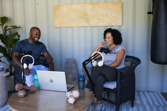 African American couple in athletic wear sitting in studio, recording with laptop, mics, dumbbells